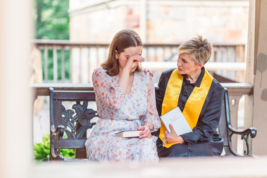 woman-in-white-floral-dress-sitting-on-bench-with-a-priest-8674118 Woman receiving emotional support from a priest on a church bench.